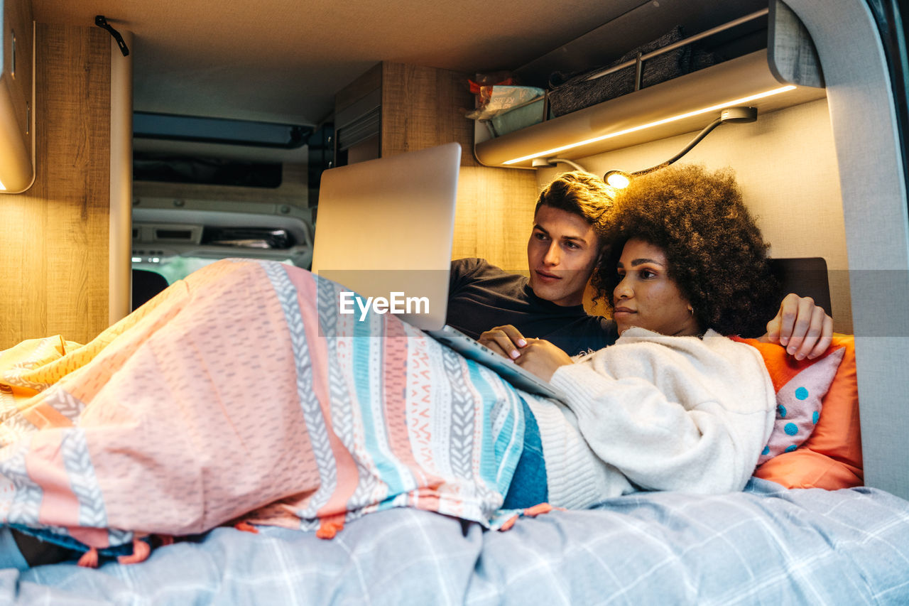 Happy romantic multiracial couple cuddling and enjoying time together while lying on bed and watching movie on laptop during travel in camper van