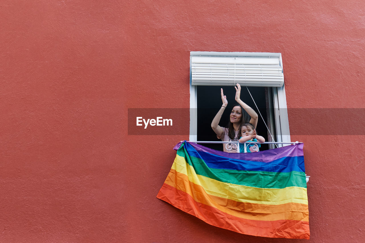Young woman in a mask clapping from the window with the gay flag on a red background