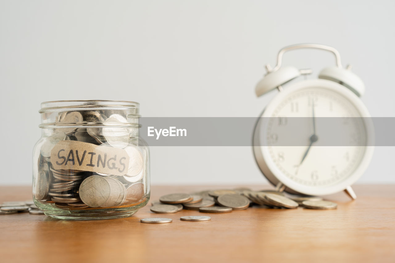Close-up of coins in jar with alarm clock on table against white wall