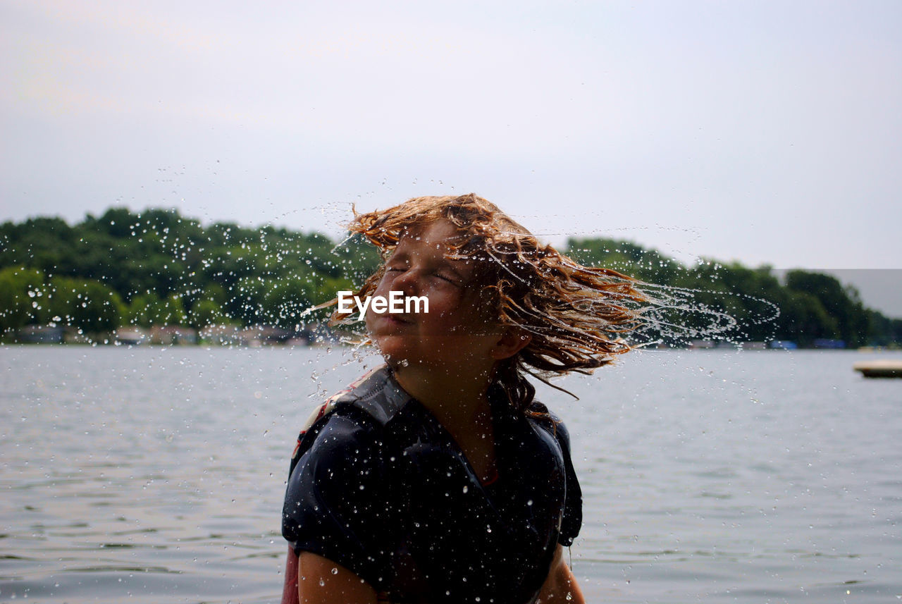 Boy tossing wet hair while standing at lake against sky