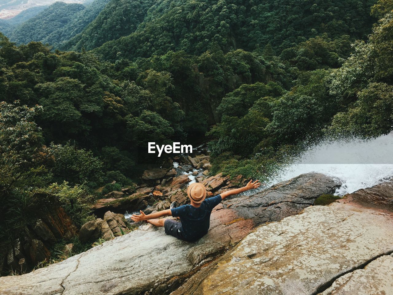 High angle view of man with arms outstretched sitting on rock formation against forest