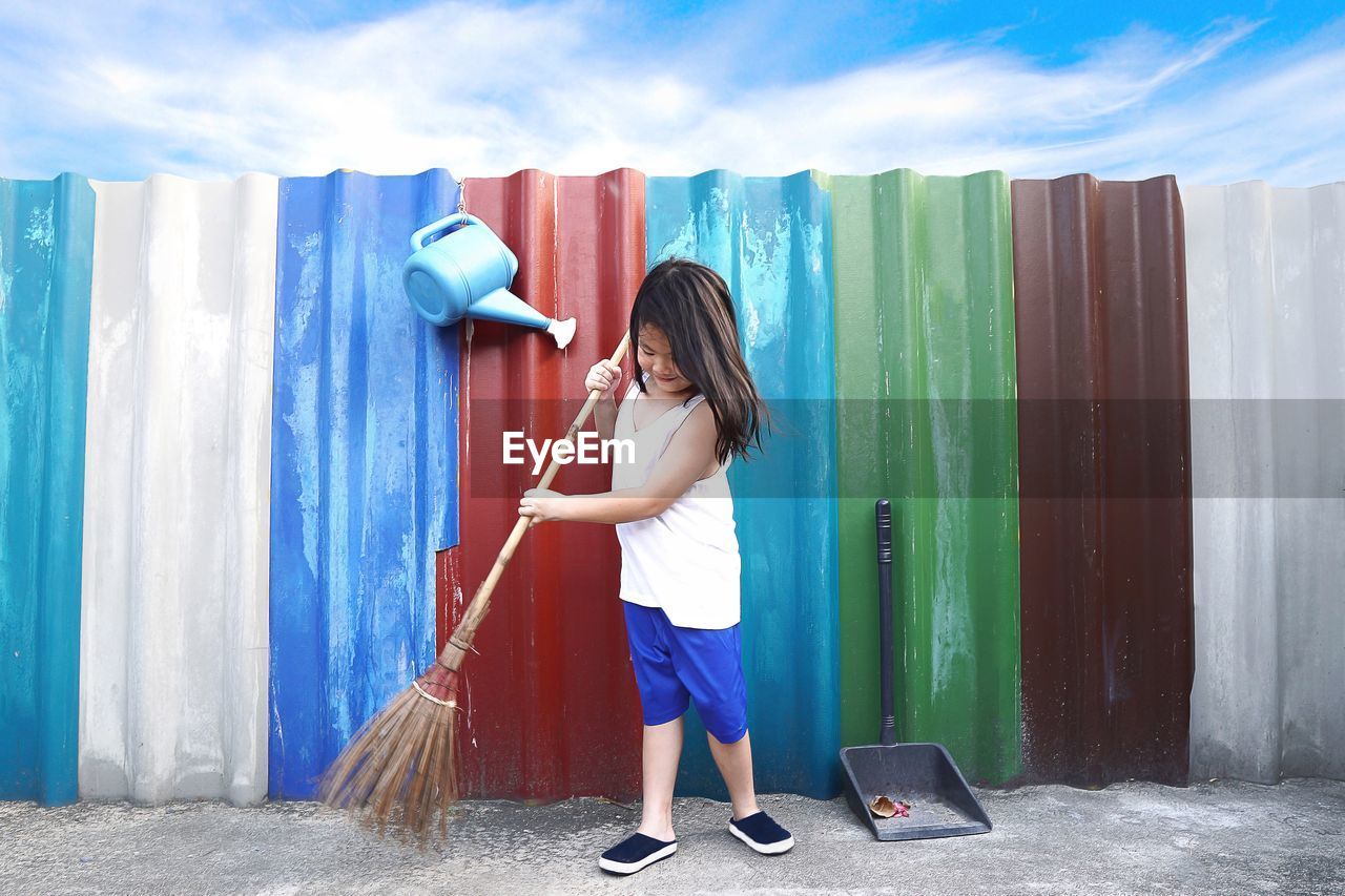 Full length of girl sweeping footpath by colorful corrugated irons