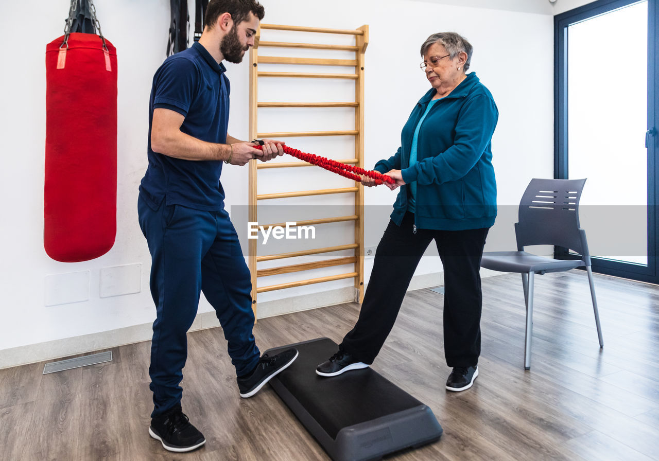 Side view of old woman and male fitness instructor standing in front on each other and pulling elastic band using step platform in gym