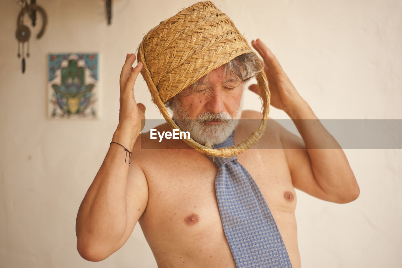 Shirtless man wearing basket on head while standing against wall