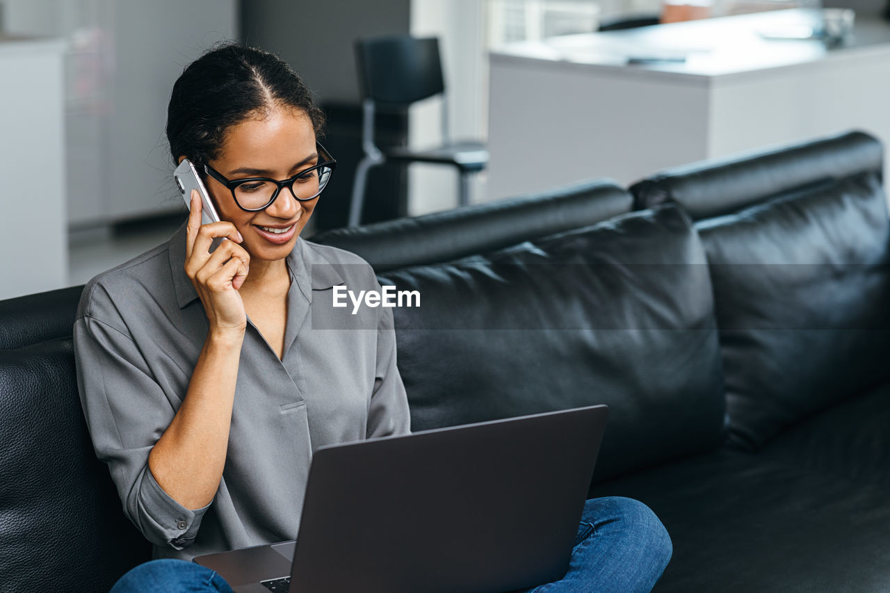 Woman talking over mobile phone while using laptop on sofa