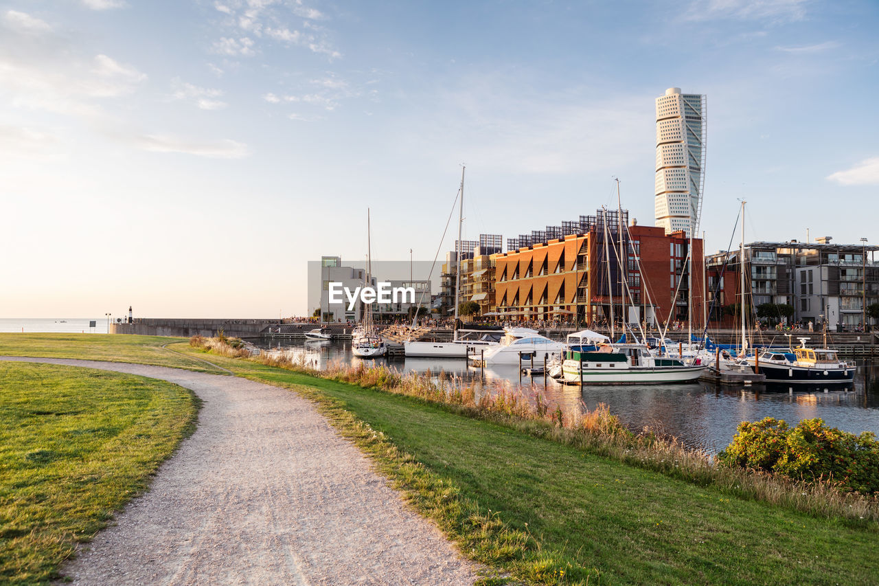 Footpath amidst grass by harbor against sky in city