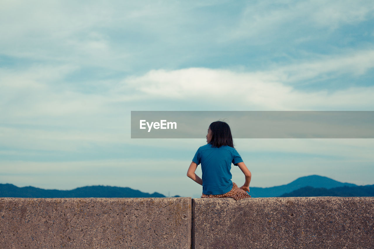 Rear view of girl sitting on retaining wall against sky