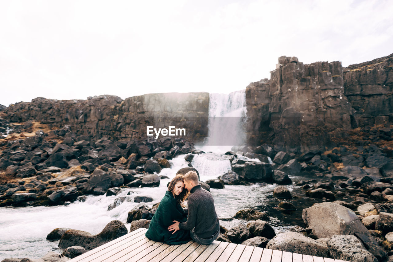 YOUNG MAN SITTING ON ROCK BY WATER AGAINST SKY