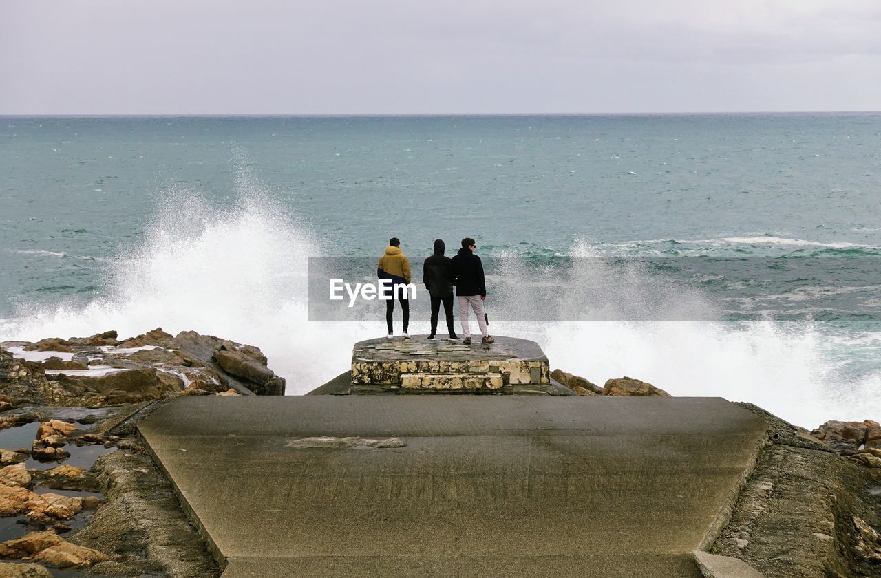 MAN STANDING ON ROCK BY SEA AGAINST SKY