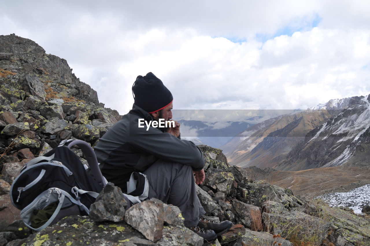 Side view of hiker sitting on mountain against cloudy sky