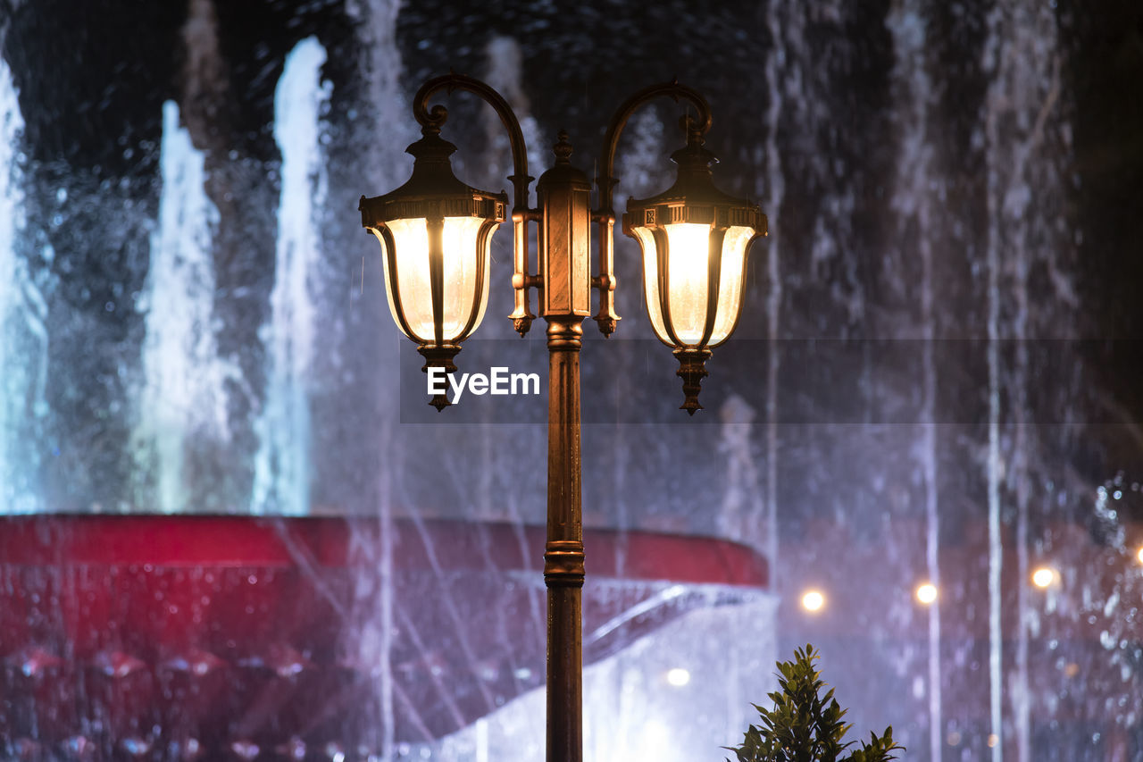 Low angle view of illuminated street lights against fountains in city at night