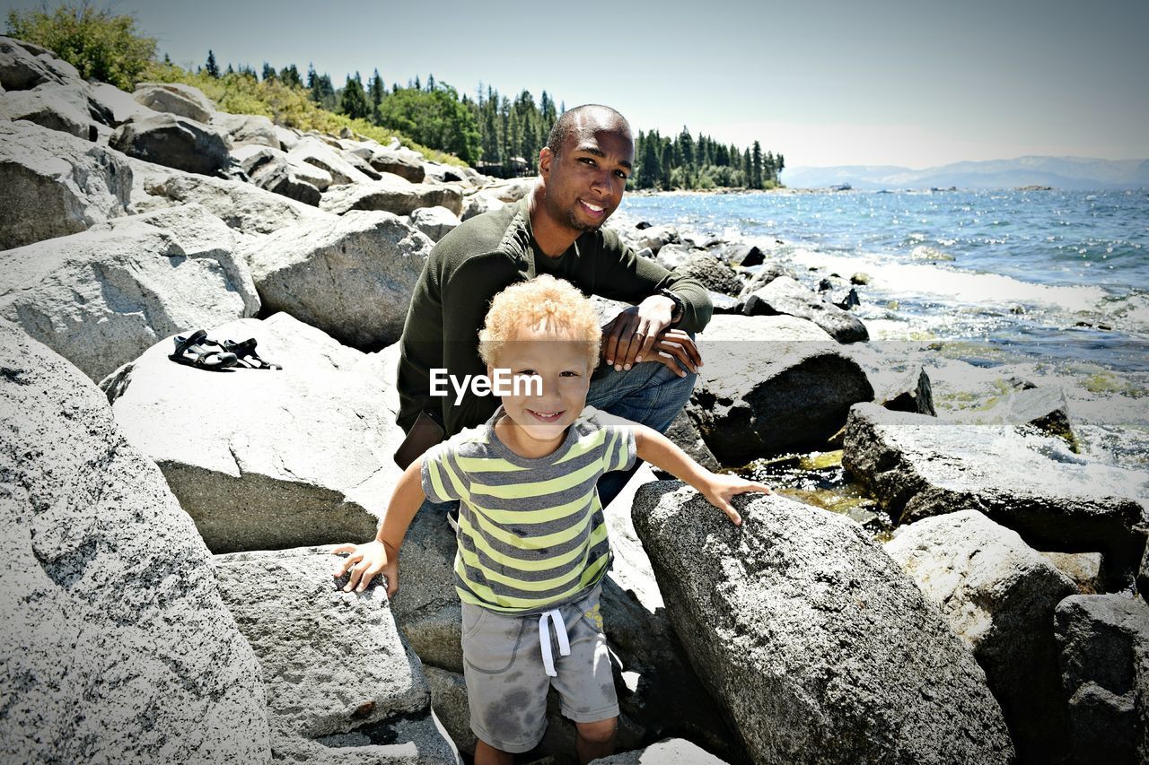 Portrait of father with son on rocks by sea during sunny day