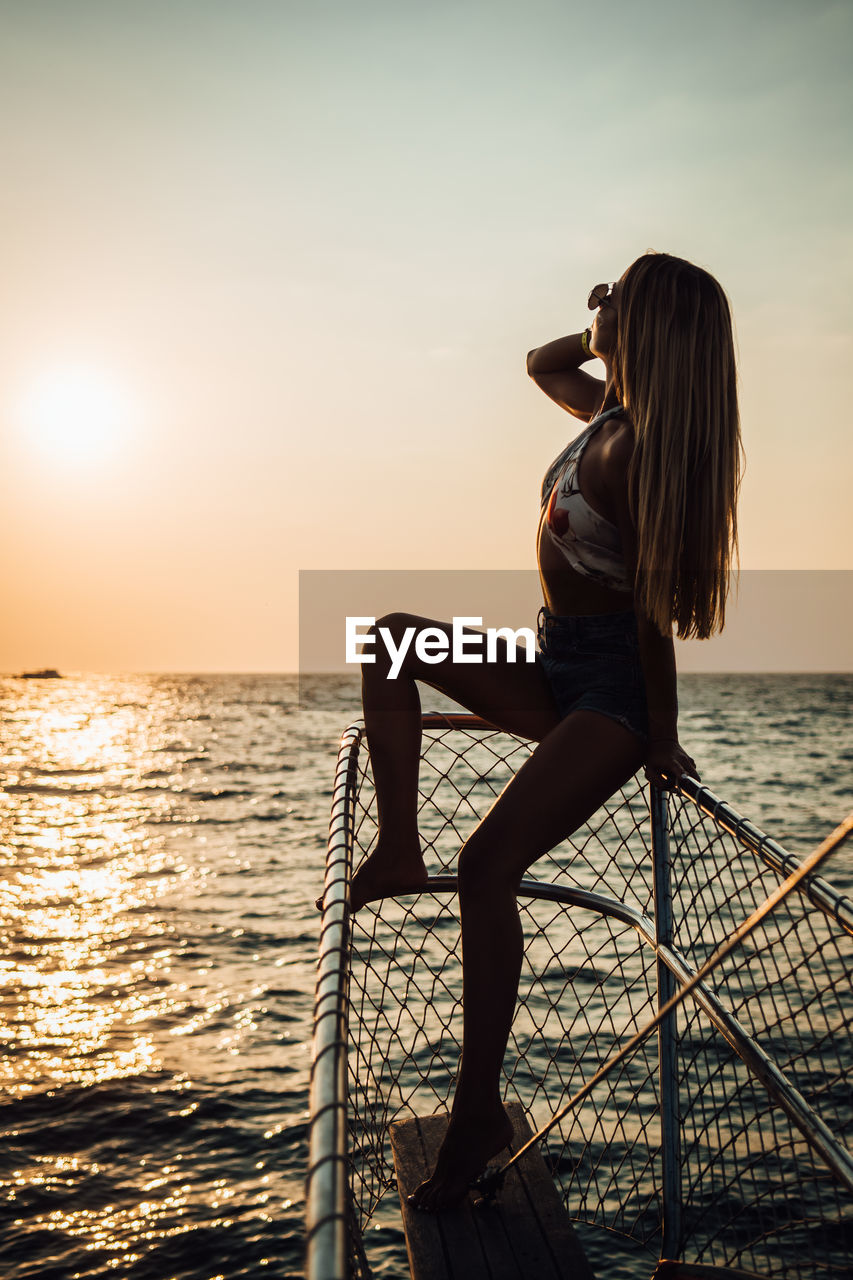 WOMAN SITTING BY SEA AGAINST SKY