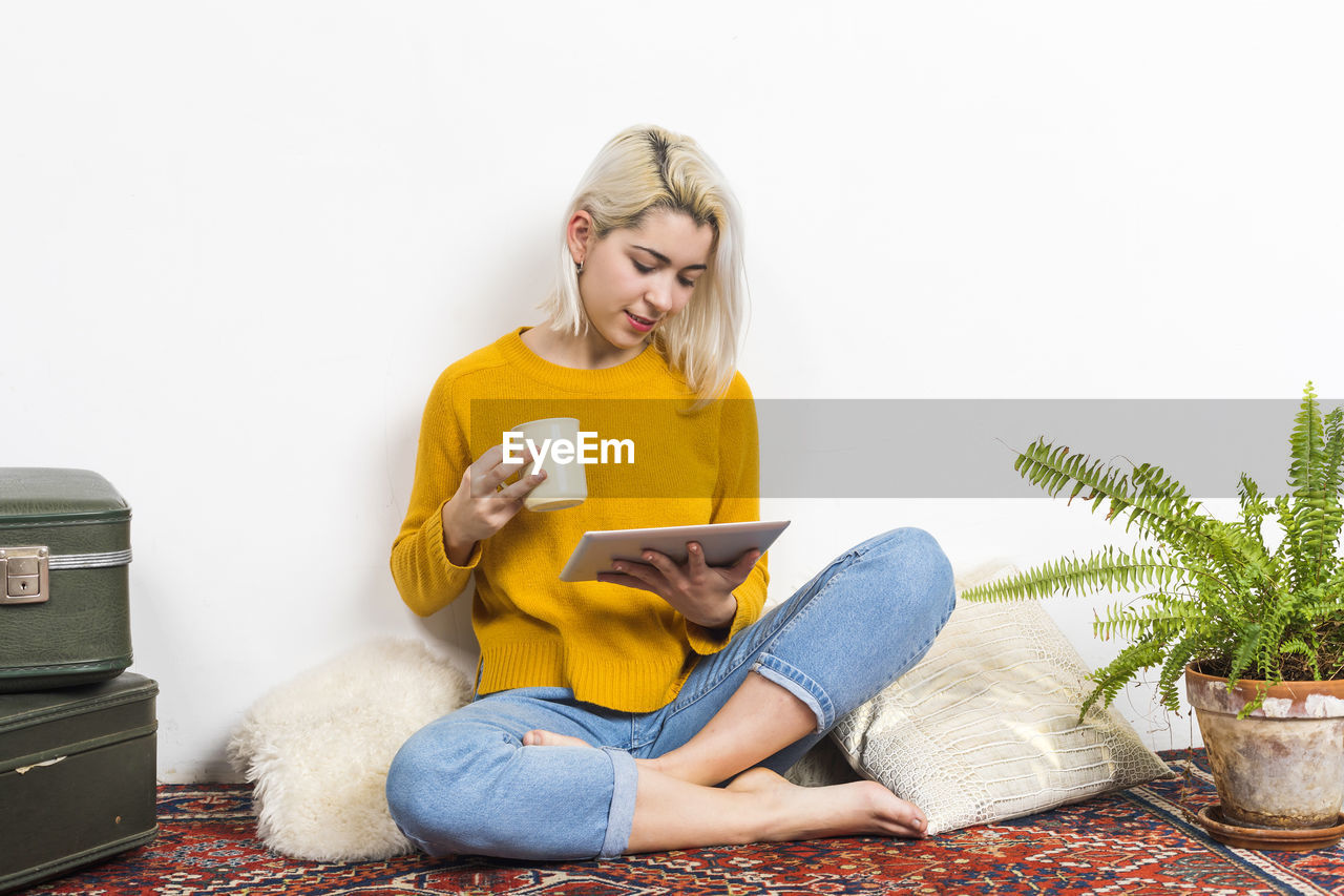 sitting, one person, adult, person, technology, women, indoors, casual clothing, blond hair, lifestyles, full length, clothing, living room, wireless technology, young adult, furniture, relaxation, communication, domestic room, home interior, yellow, leisure activity, front view, computer, domestic life, hairstyle, sofa, music, cross-legged, jeans, looking, plant, footwear, reading, digital tablet, photo shoot, holding, arts culture and entertainment, houseplant, long hair, smiling, female, publication, looking down, internet, eyeglasses, glasses, nature, emotion