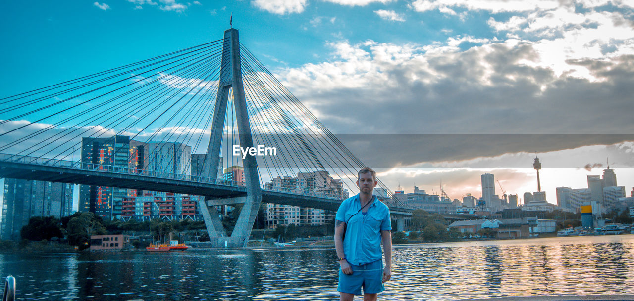 Portrait of man standing by river against suspension bridge