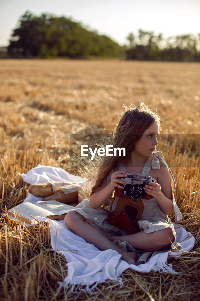 Girl child in a dress sitting on a mown field with a camera on a blanket with bread and a book