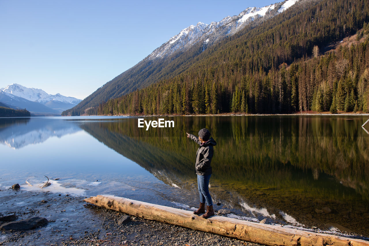 Side view of young woman walking on log at lakeshore against mountain