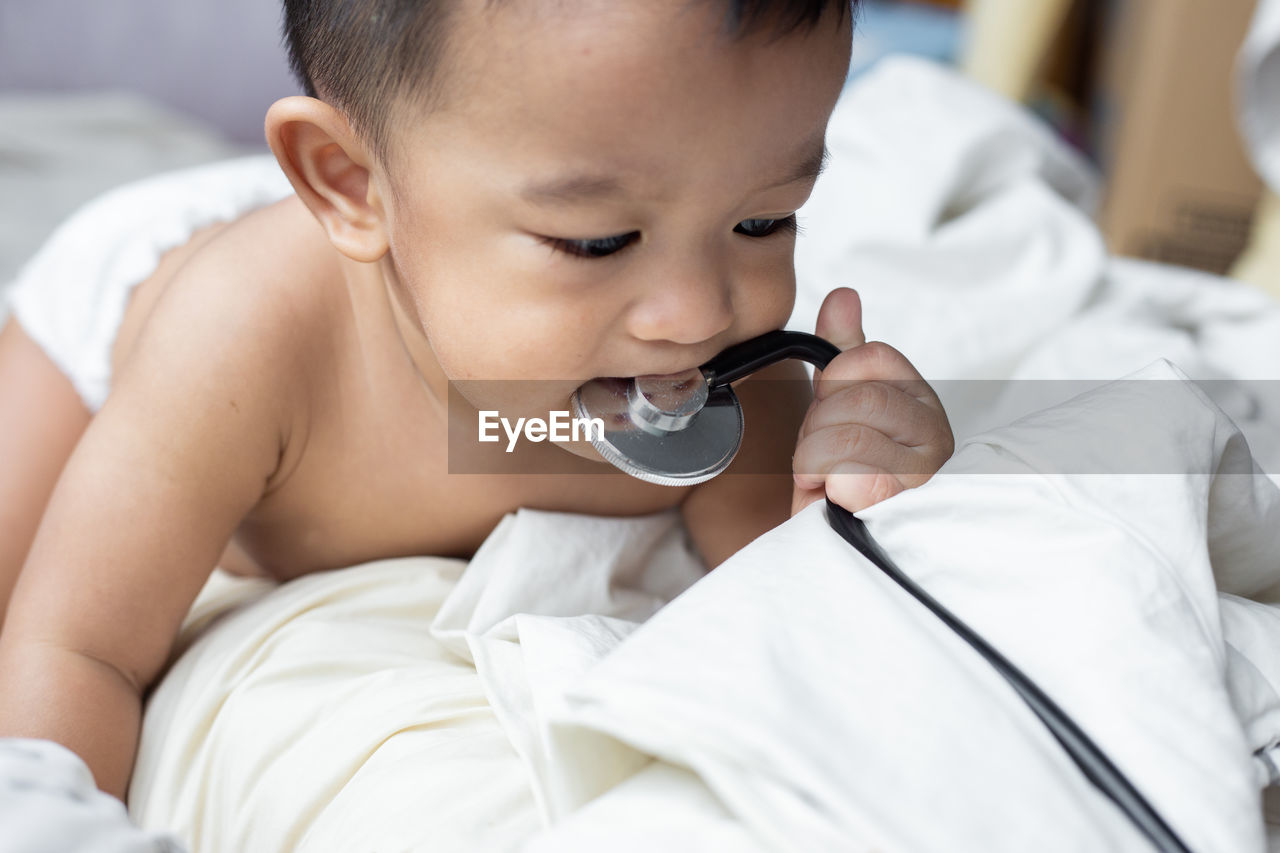 Cute baby boy biting stethoscope while lying on bed at home