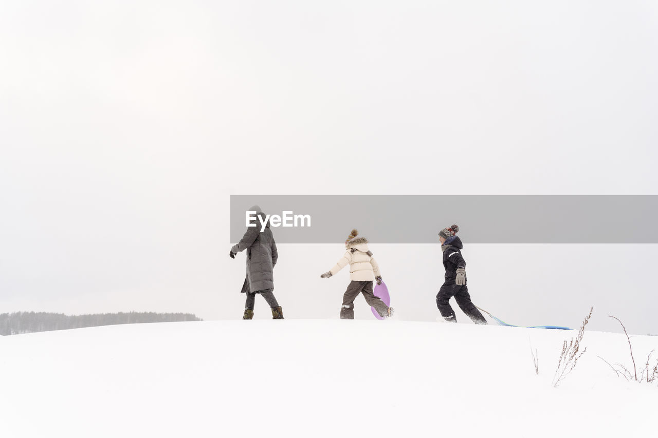 Father and children walking with sleds on snow covered land against sky