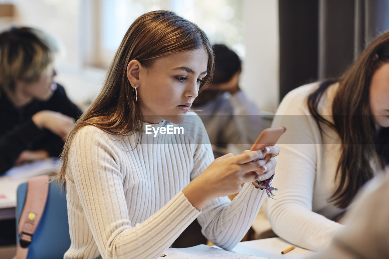 Teenage girl with brown hair using smart phone sitting by female friend at desk in classroom