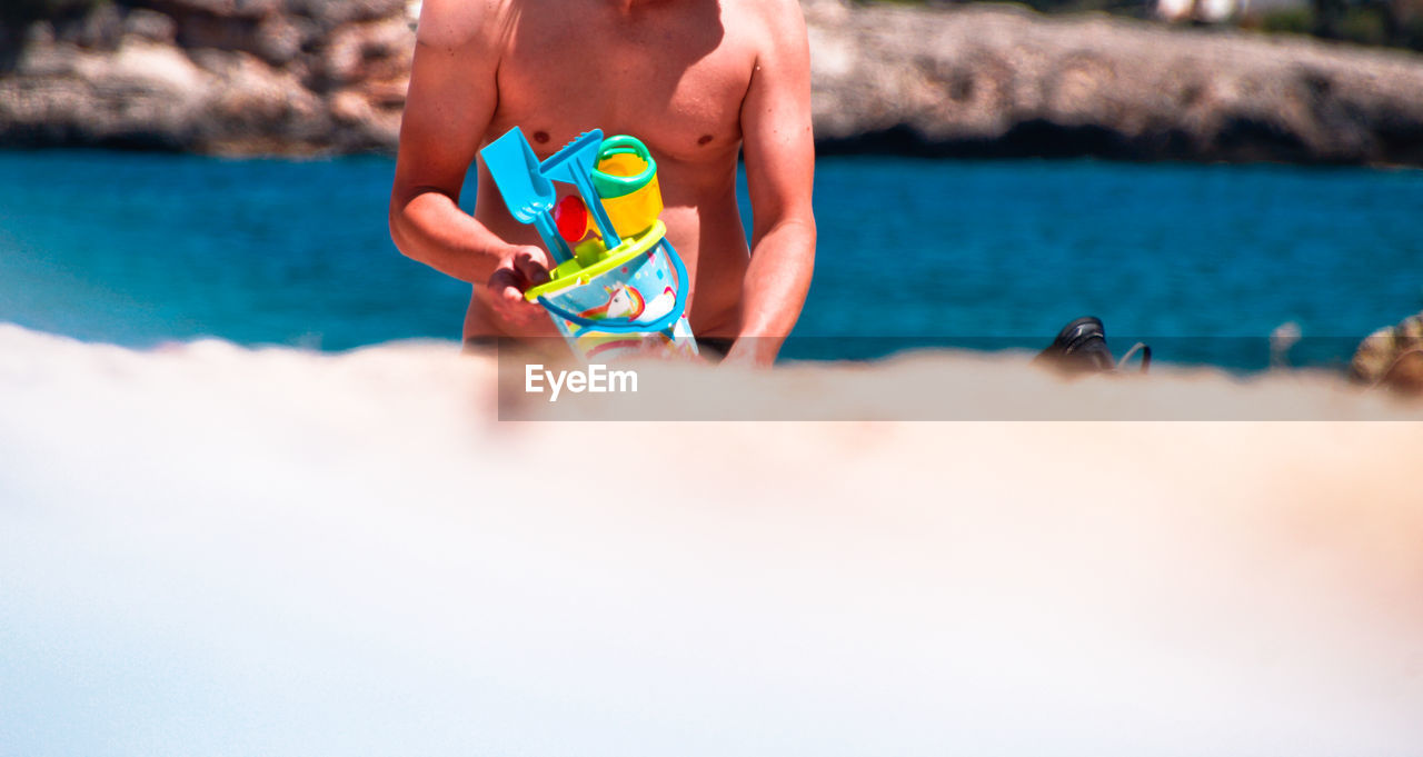 Midsection of shirtless man holding toys at beach