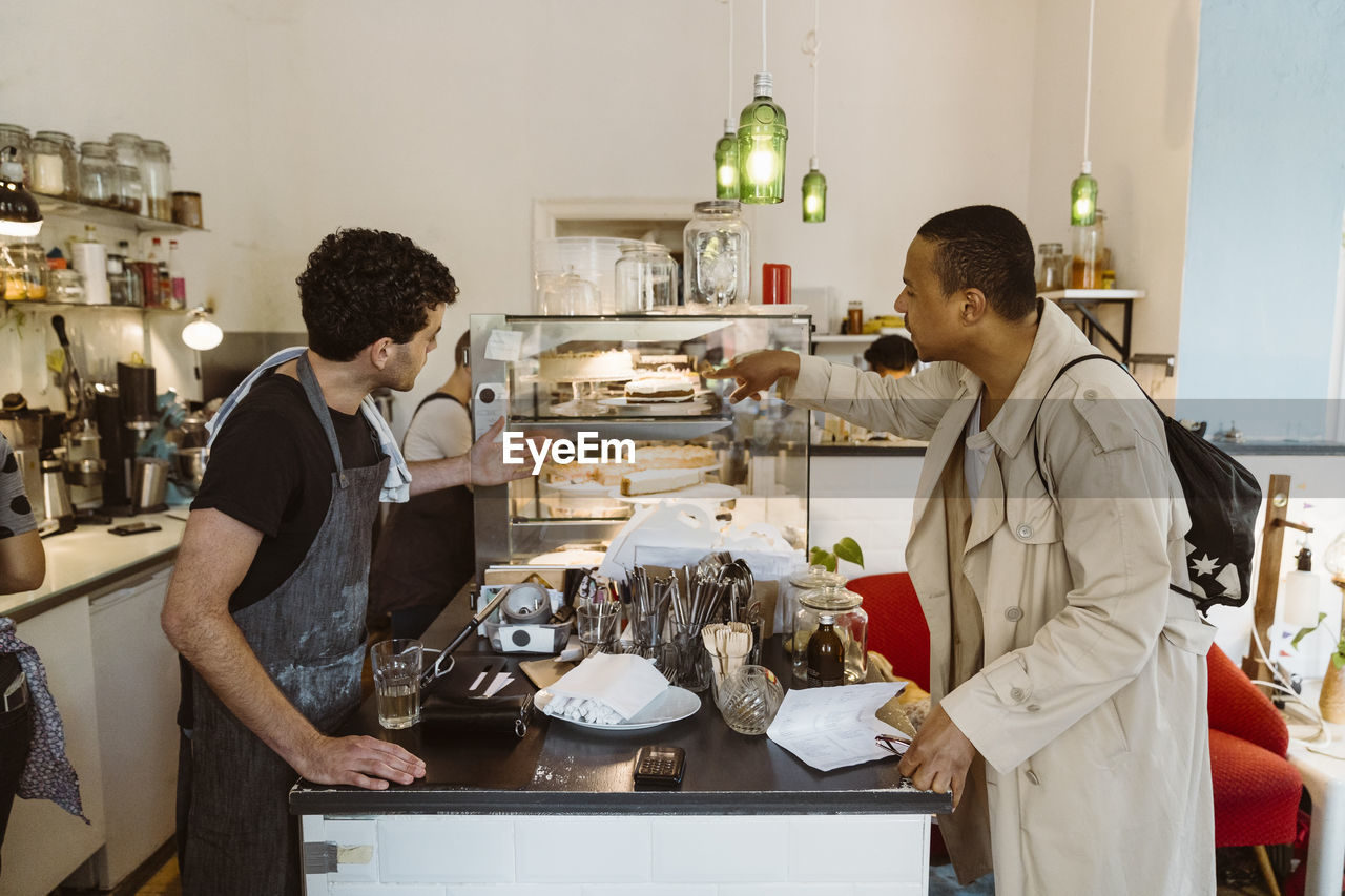 Side view of male cafe owner suggesting food to customer at counter