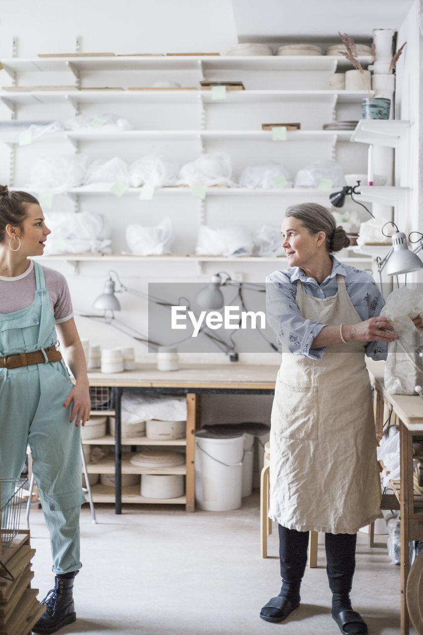 Senior female potter working with young employee against shelves at workshop