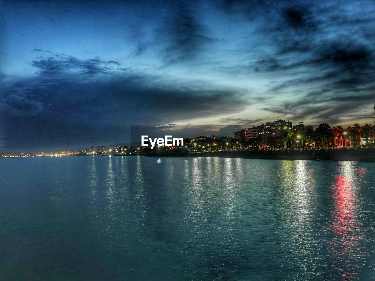 SEA BY ILLUMINATED BUILDINGS AGAINST SKY AT DUSK