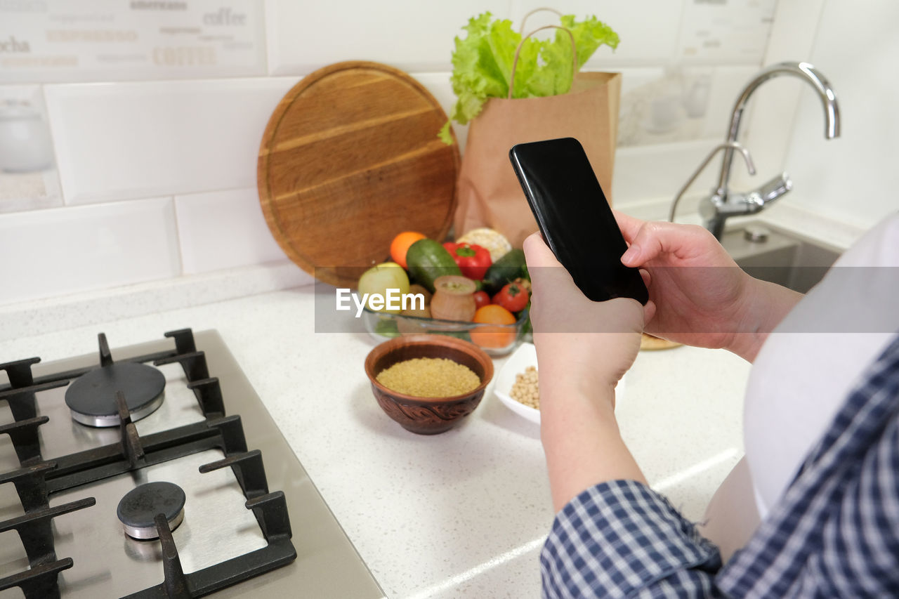Female hand holding cell phone with empty screen on background kitchen table with grocery