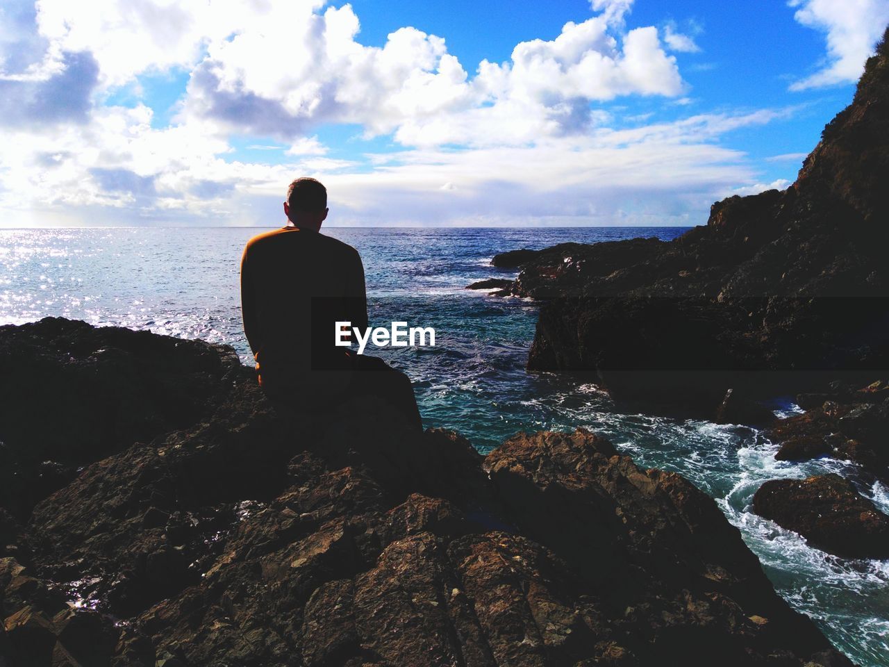 Rear view of man sitting on rock over sea against sky