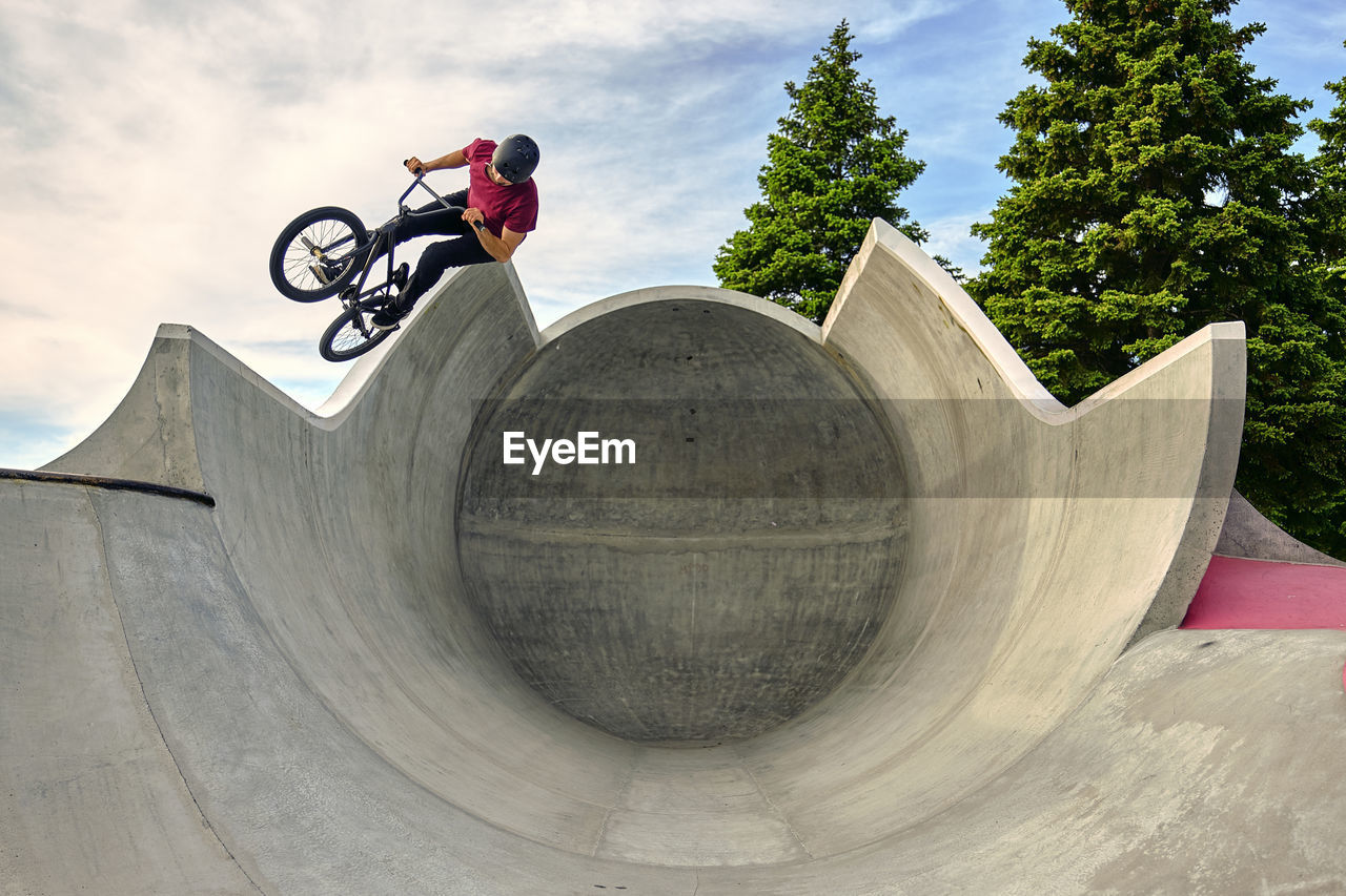 Male rider with bmx bike jumping on concrete ramp against sky at skateboard park