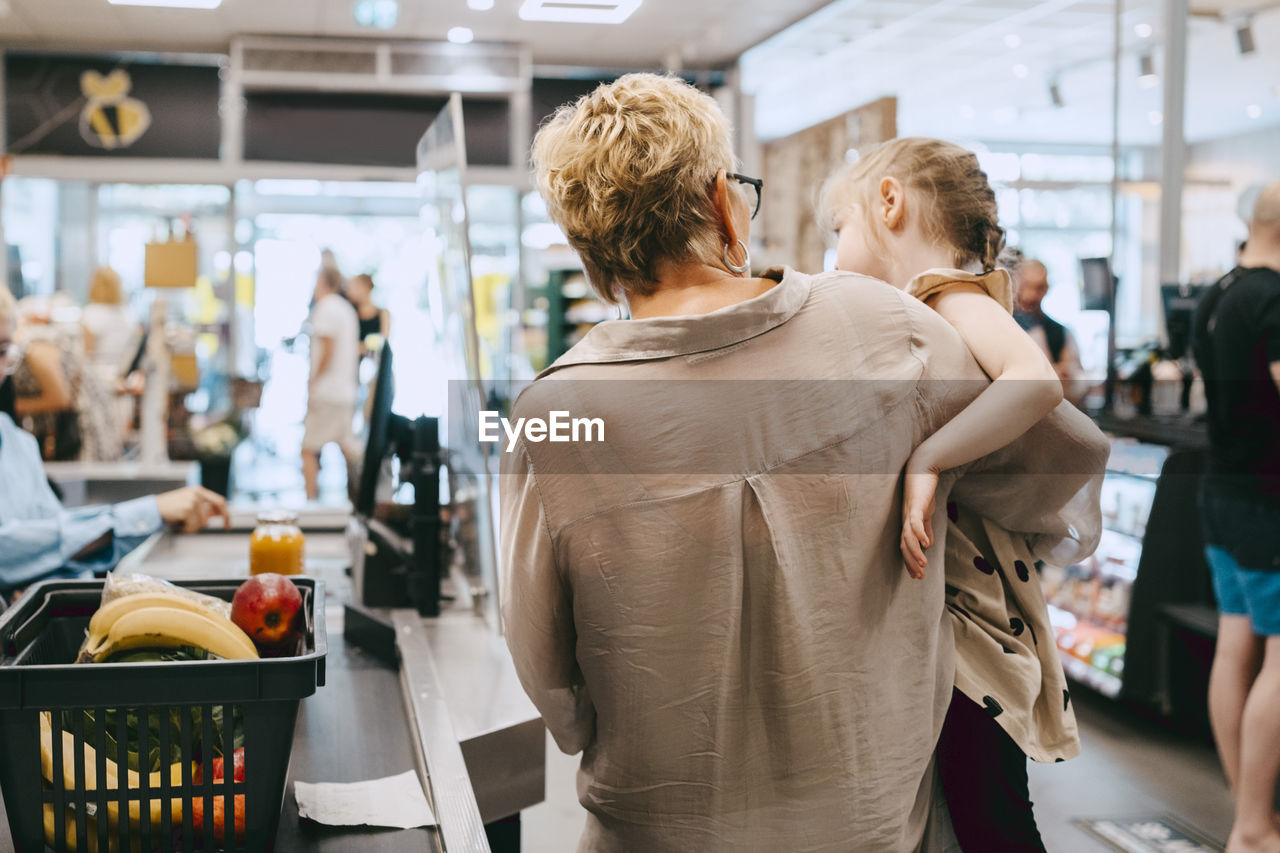 Grandmother carrying granddaughter while standing at checkout counter in supermarket