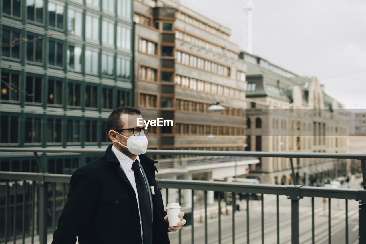 Male entrepreneur with coffee cup by railing in city during pandemic