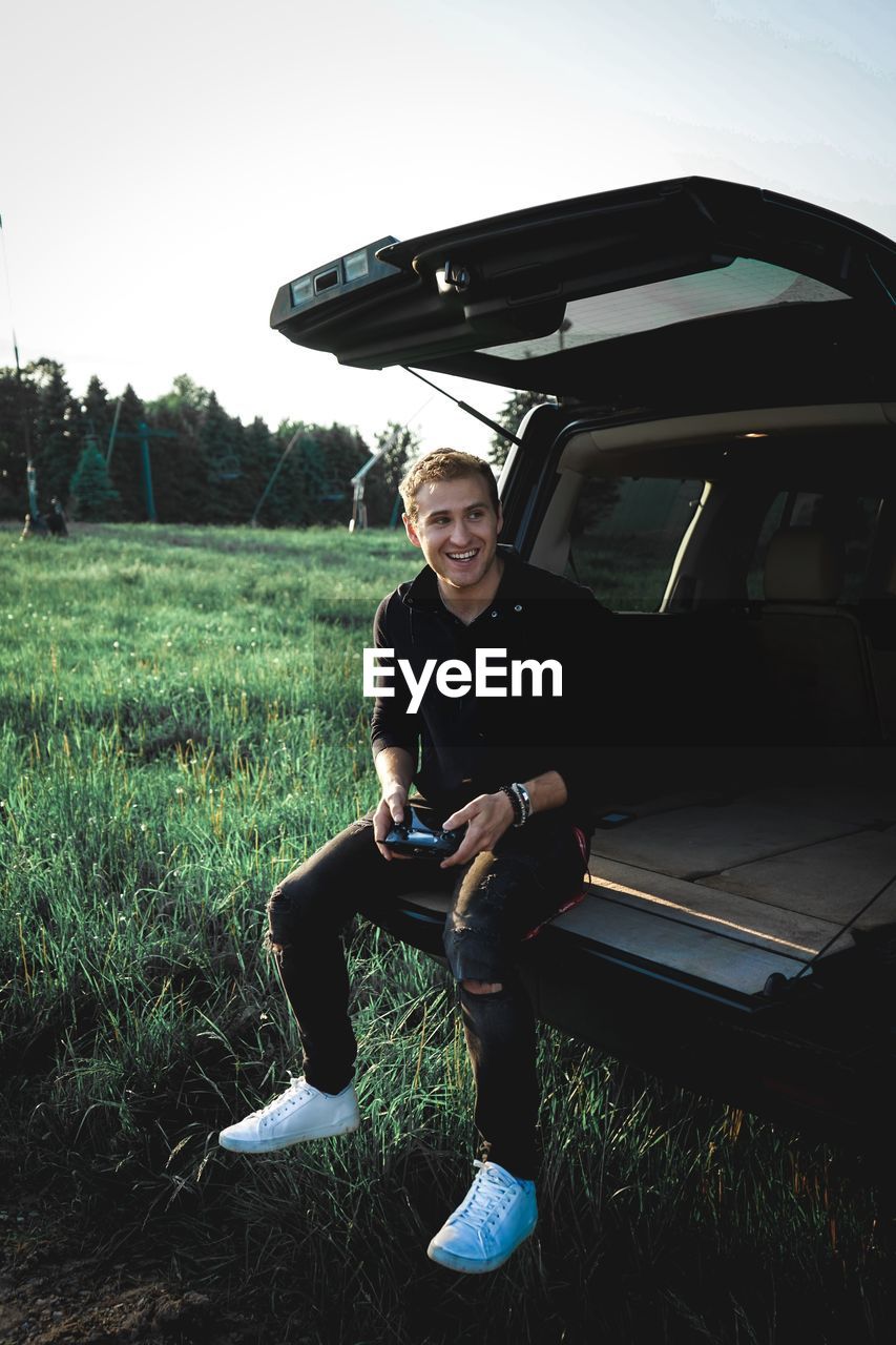 Young man smiling while sitting in car trunk on grassy field