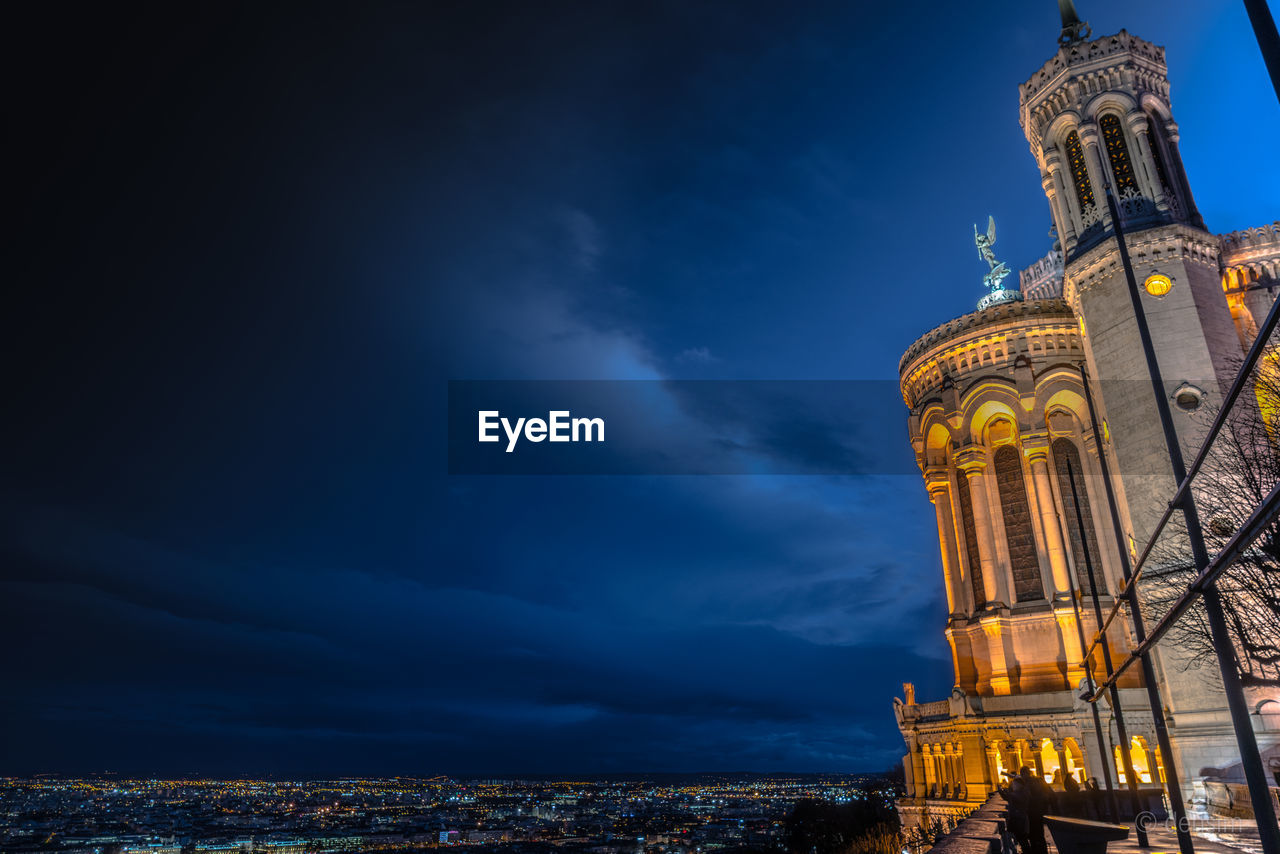 Historical building with illuminated city in background against cloudy sky