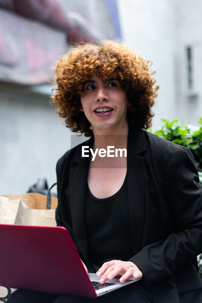 A non-binary person looking to the right working on the laptop and a  shopping bag in the background