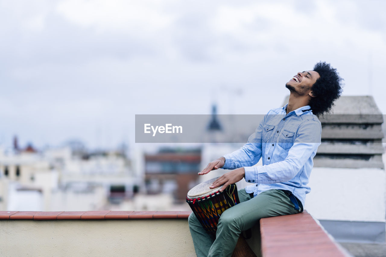 Happy young man playing drum while sitting on terrace against sky