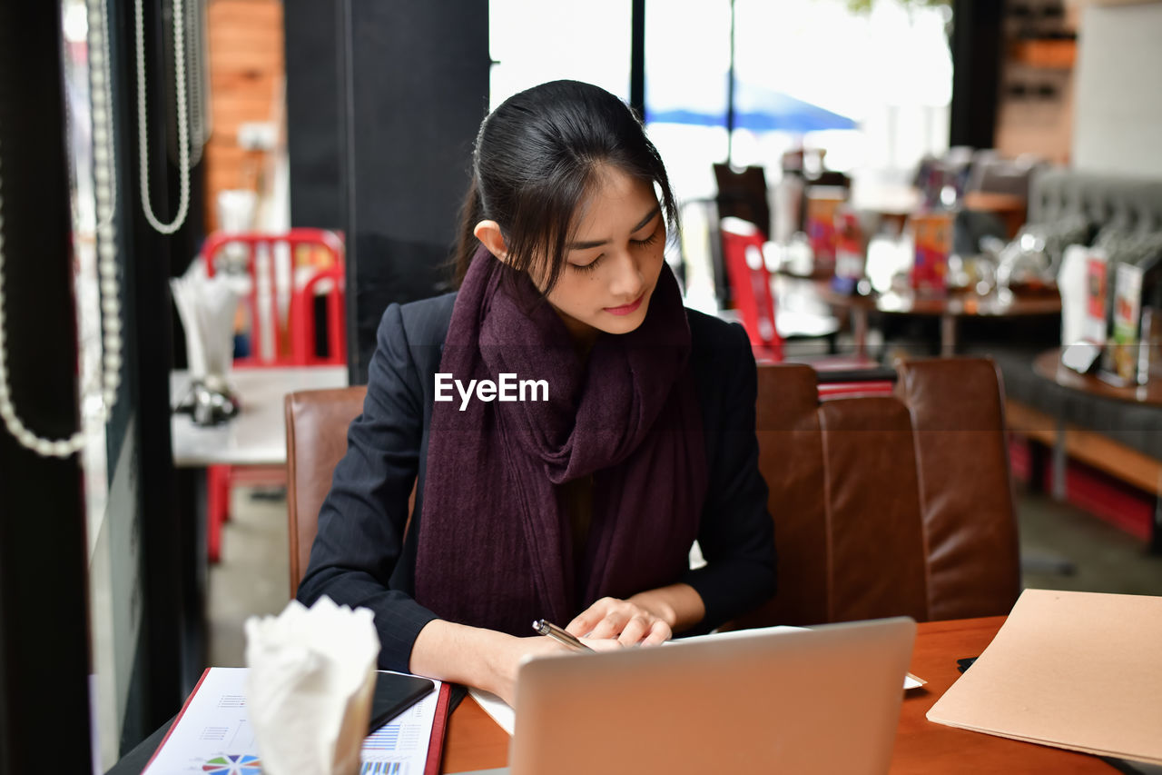 Young woman writing in book by laptop on table