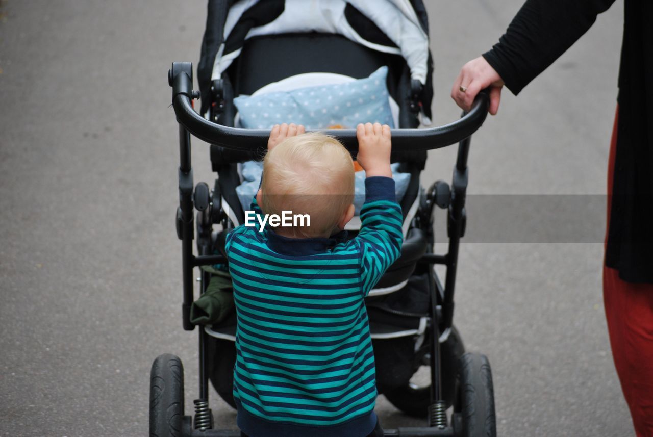 Cropped image of person walking with boy pushing baby stroller on footpath