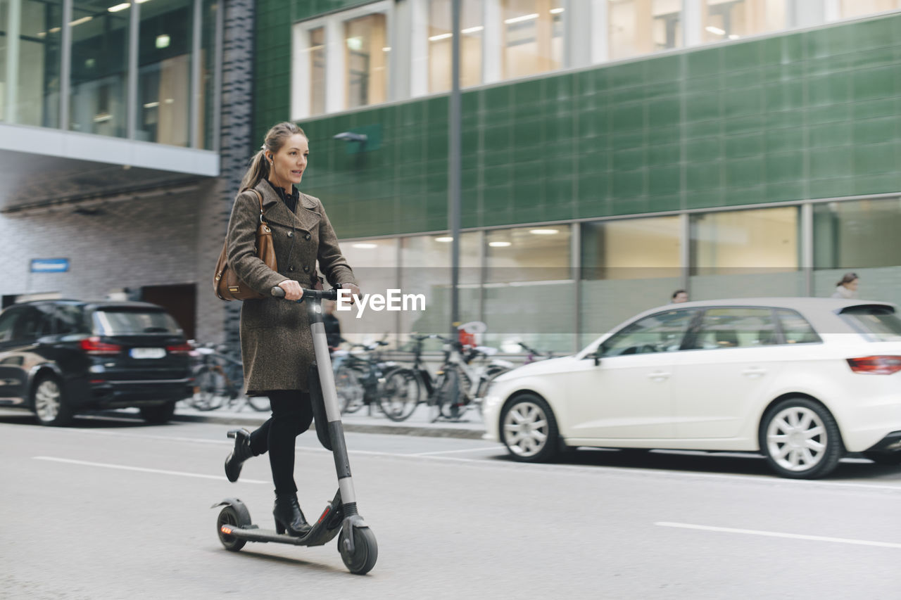 Full length of confident businesswoman riding electric push scooter on street in city