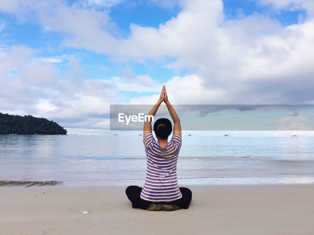 Rear view of woman meditating while sitting on beach against cloudy sky