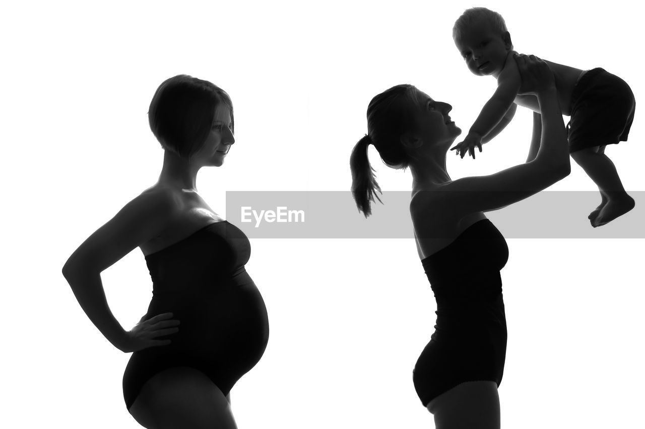 Side view of pregnant woman standing with sister holding toddler son against white background