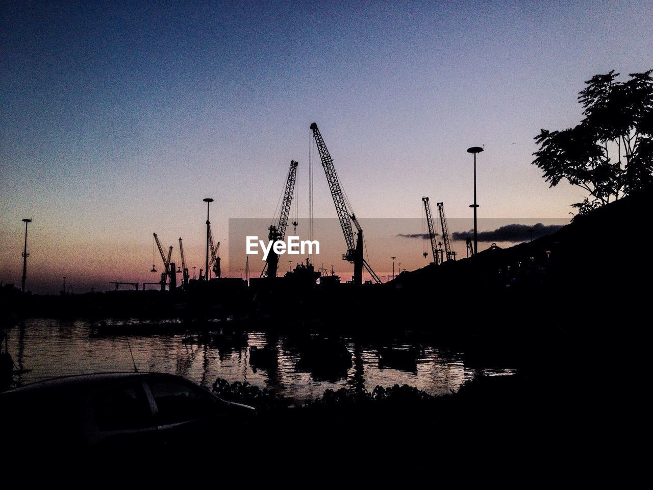 Silhouette cranes at dock by sea against sky during sunset