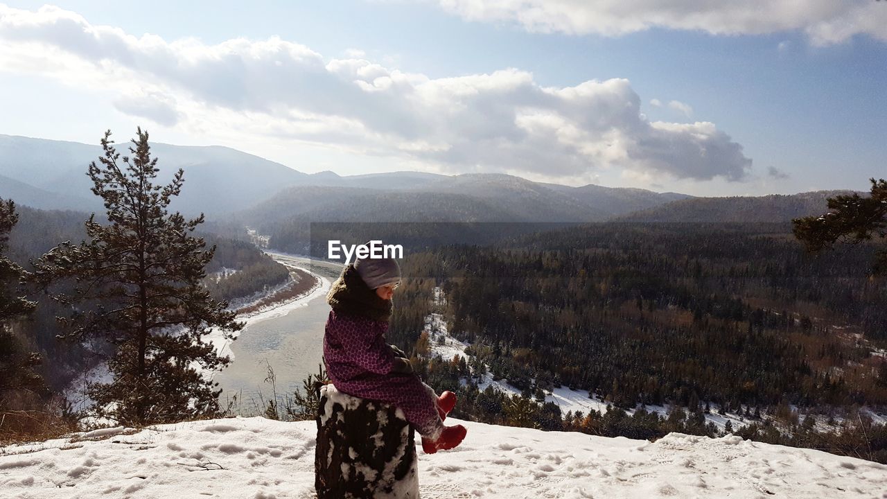 Girl wearing warm clothing while sitting on snowcapped mountain during winter