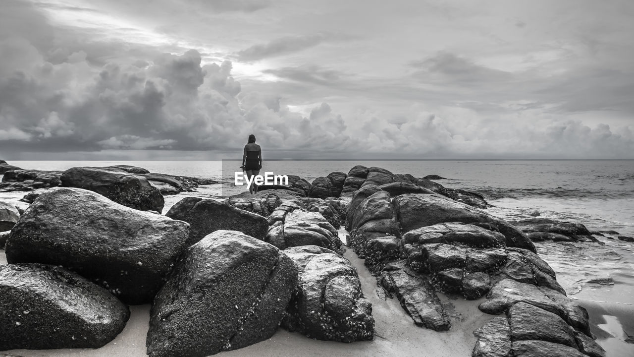 Rear view of woman standing on rocks at beach against sky
