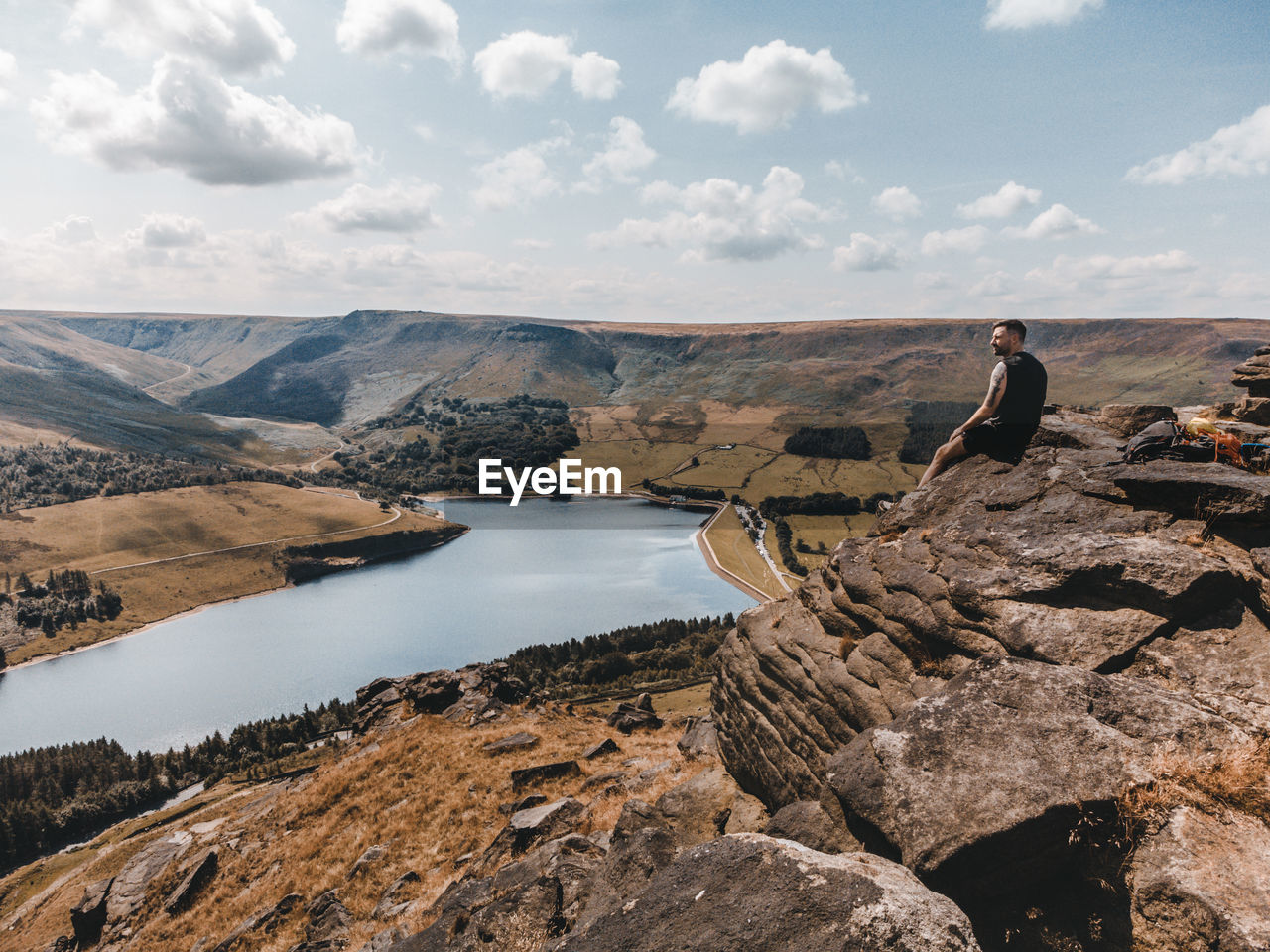 Lone hiker sat on cliff edge overlooking lake shot by a drone