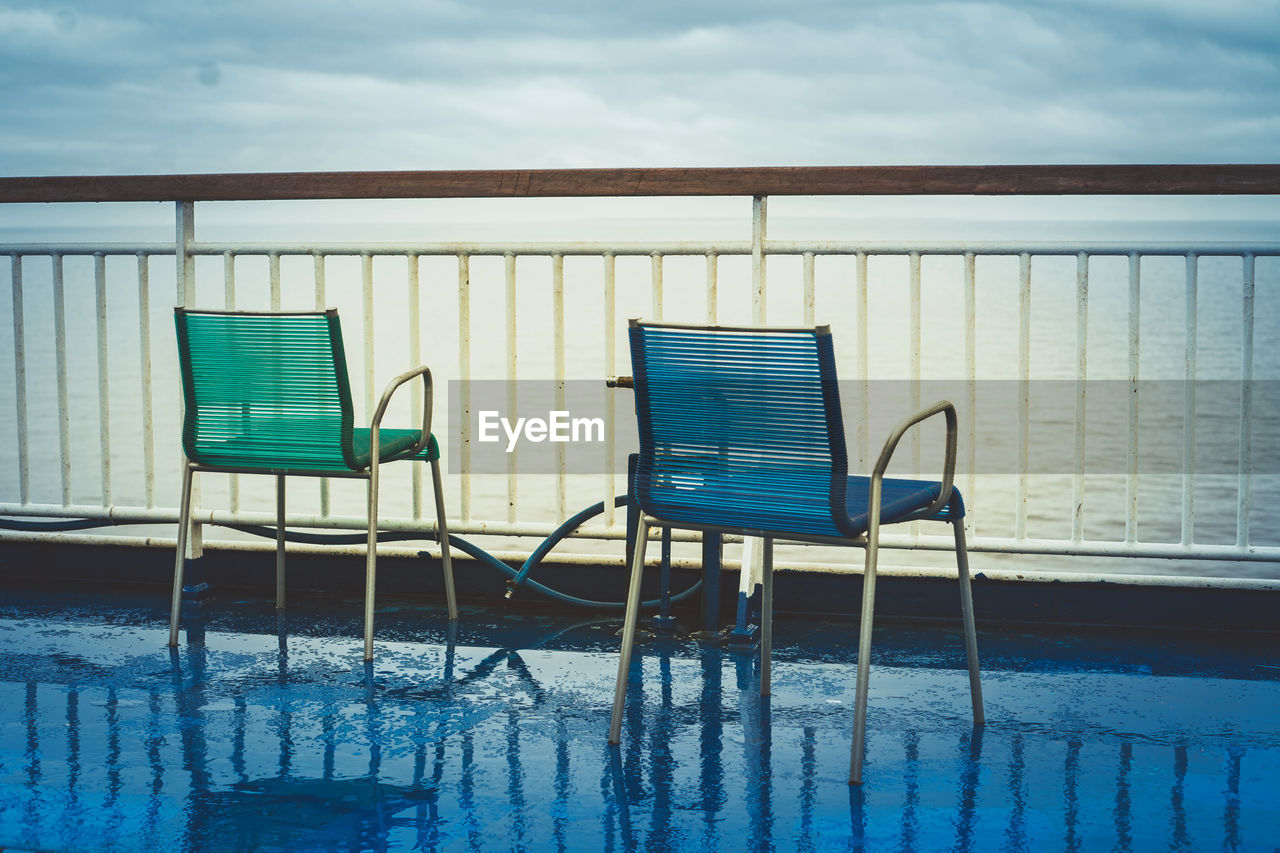 Empty chairs on pier by sea against cloudy sky