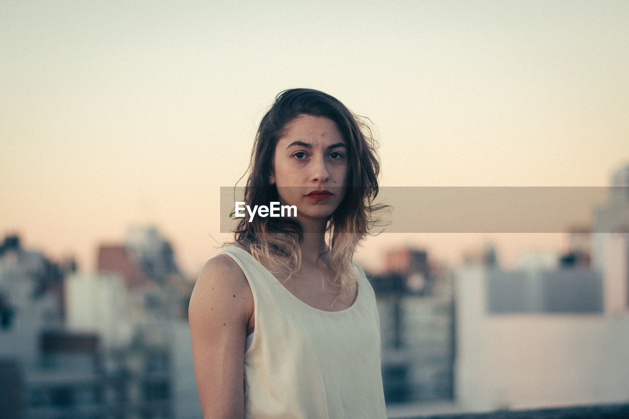 Portrait of young woman standing against clear sky in city during sunset