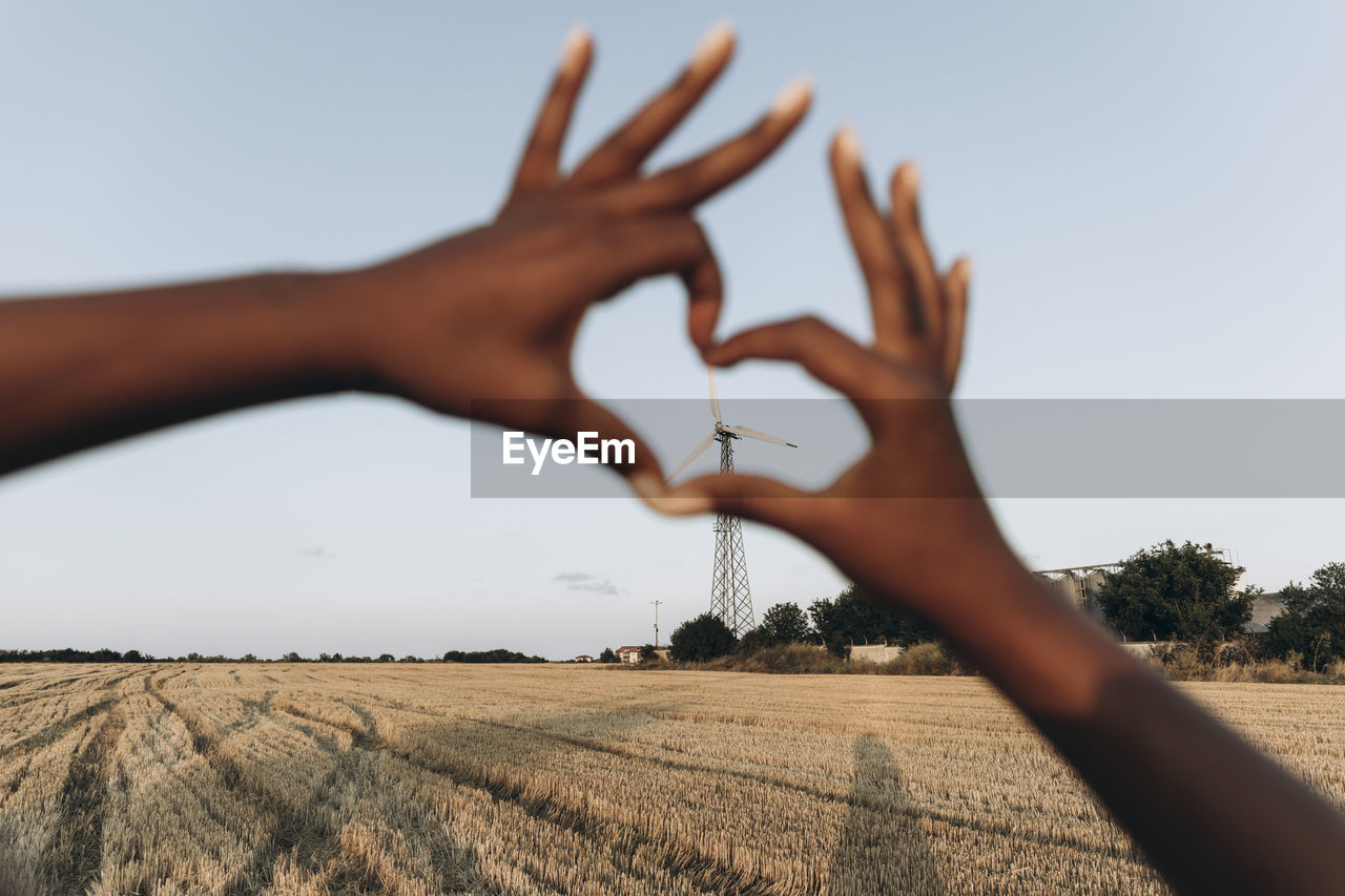 Woman making heart shape with hands in front of sky at farm