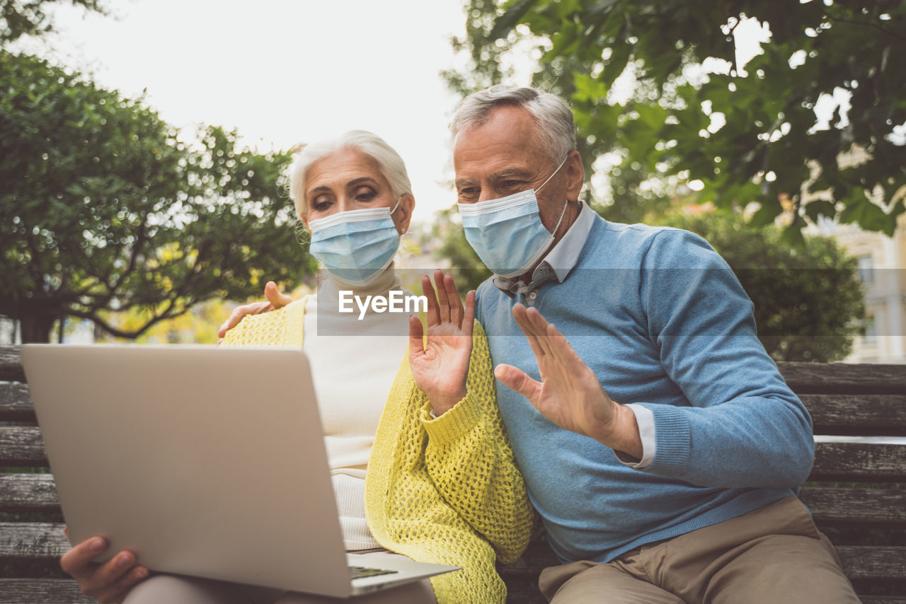 Smiling senior couple wearing mask video calling sitting at park